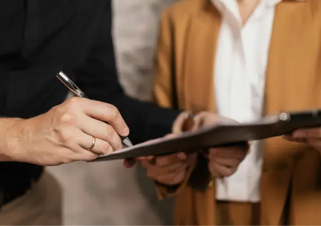 Dos personas están de pie una al lado de la otra; una sostiene una carpeta con documentos mientras la otra firma contratos con una pluma. No se les ve el rostro.