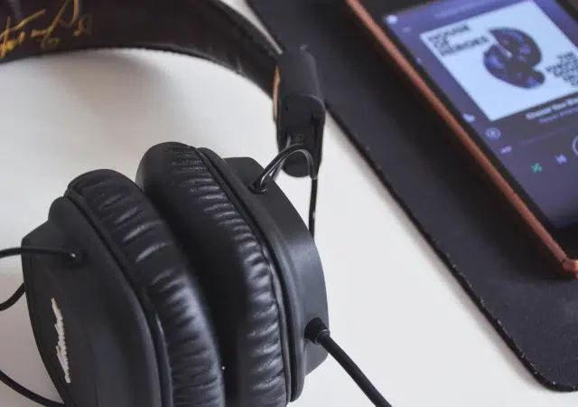Unos audífonos negros de diadema están colocados junto a un smartphone que muestra playlists de Spotify sobre un mousepad negro.