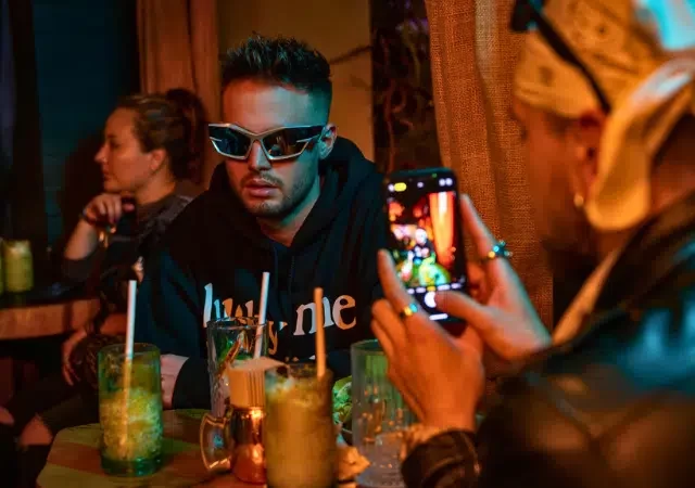 Un hombre con gafas de sol se sienta a una mesa con bebidas, mientras otra persona le toma una foto con un smartphone—quizás capturando contenido sobre cómo ganar dinero en TikTok en un ambiente tenuemente iluminado.