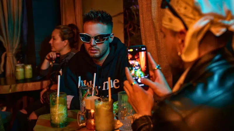 Un hombre con gafas de sol se sienta a una mesa con bebidas, mientras otra persona le toma una foto con un smartphone—quizás capturando contenido sobre cómo ganar dinero en TikTok en un ambiente tenuemente iluminado.