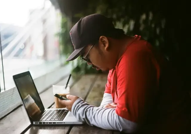 Un hombre con camiseta y gorra rojas se sienta en una mesa de madera con una laptop y una bebida, mirando su smartphone cerca de una gran ventana, probablemente leyendo newsletters de la industria musical.
