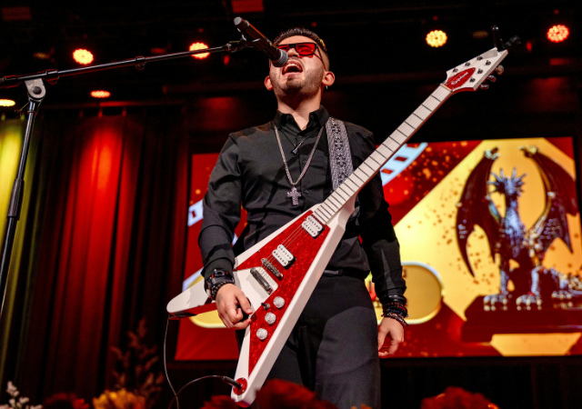 Un músico vestido de negro y usando gafas oscuras toca una guitarra eléctrica roja y canta en el escenario, con una estatua de dragón proyectada en pantalla detrás de él durante los Latin GRAMMY Awards.