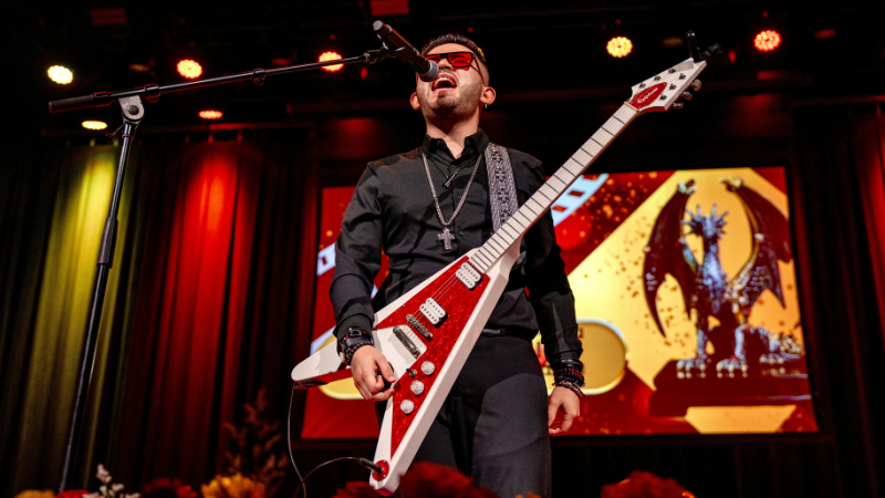 Un músico vestido de negro y usando gafas oscuras toca una guitarra eléctrica roja y canta en el escenario, con una estatua de dragón proyectada en pantalla detrás de él durante los Latin GRAMMY Awards.