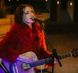 Una mujer con un abrigo de piel rojo toca una guitarra acústica y canta al micrófono sobre el escenario, con un teclado de fondo, ofreciendo una interpretación digna de los GRAMMY.
