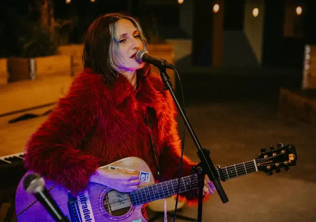 Una mujer con un abrigo de piel rojo toca una guitarra acústica y canta al micrófono sobre el escenario, con un teclado de fondo, ofreciendo una interpretación digna de los GRAMMY.