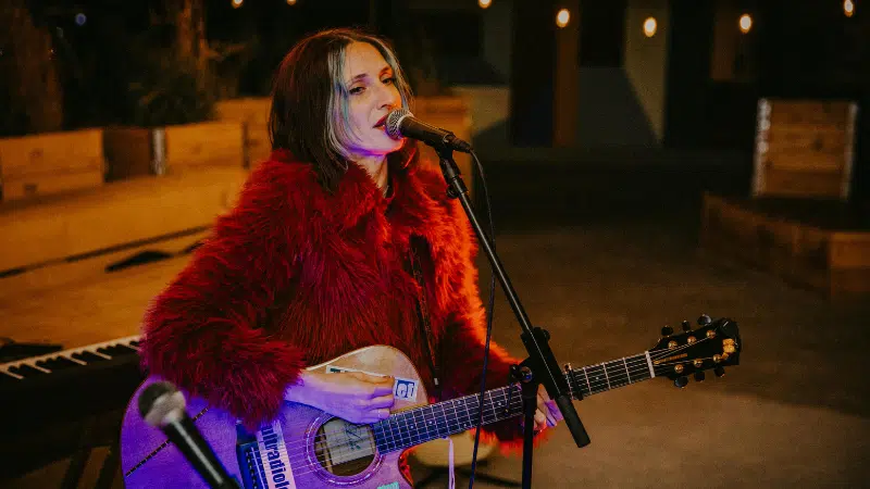 Una mujer con un abrigo de piel rojo toca una guitarra acústica y canta al micrófono sobre el escenario, con un teclado de fondo, ofreciendo una interpretación digna de los GRAMMY.