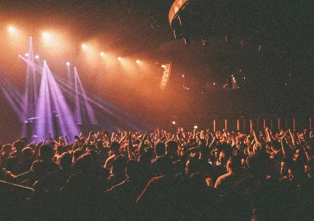 Una gran multitud se reúne con las manos en alto en un concierto o evento en vivo, iluminada por vibrantes luces moradas y naranjas del escenario, con una energía eléctrica palpable en el ambiente.