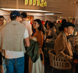 Un grupo de personas socializa y conversa en un evento en interior, algunas de pie y otras sentadas alrededor de mesas bajo una iluminación cálida, mientras suena música latina animada de fondo.