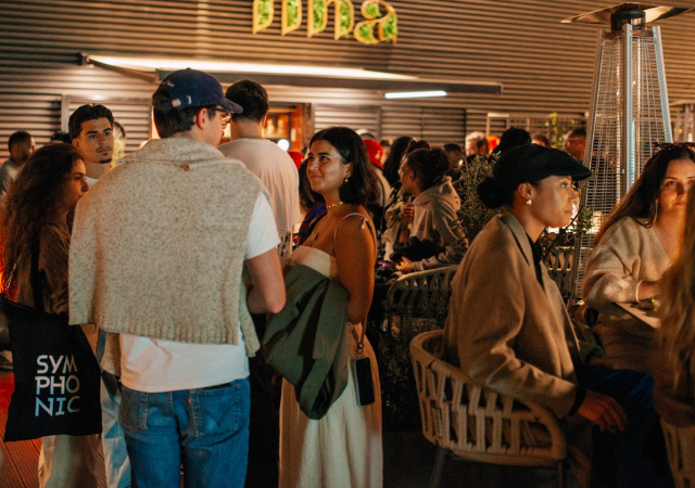 Un grupo de personas socializa y conversa en un evento en interior, algunas de pie y otras sentadas alrededor de mesas bajo una iluminación cálida, mientras suena música latina animada de fondo.
