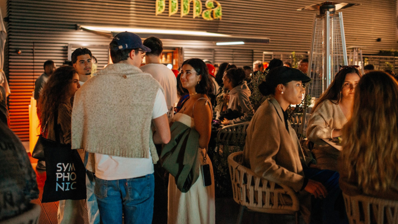 Un grupo de personas socializa y conversa en un evento en interior, algunas de pie y otras sentadas alrededor de mesas bajo una iluminación cálida, mientras suena música latina animada de fondo.
