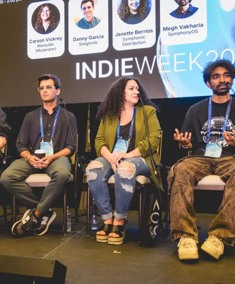Cuatro panelistas sentados en el escenario durante una conferencia de Indie Week, con gafetes y una pantalla detrás mostrando sus nombres y roles.