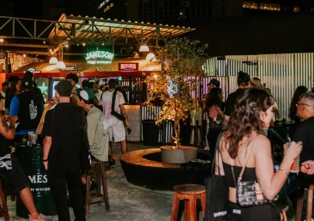 Un grupo de personas socializa en un bar al aire libre por la noche, con bebidas en la mano y letreros iluminados de fondo, evocando los encuentros animados que suelen darse después de las conferencias musicales.