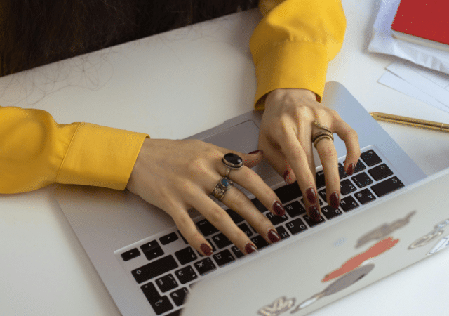 Una persona con una camiseta amarilla escribe en una laptop llena de calcomanías, rodeada de documentos, una taza y herramientas de escritura sobre una mesa blanca. Quizás está redactando un correo electrónico en español o tomando notas para su próximo gran proyecto.