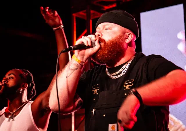 Dos hombres se presentan en el escenario con micrófonos, uno con gorro negro y overol, el otro con camiseta sin mangas blanca, frente a un fondo de luces coloridas. Su energía y pasión musical brillan mientras entretienen al público, esperando que su arte consiga el financiamiento necesario para shows más grandes.