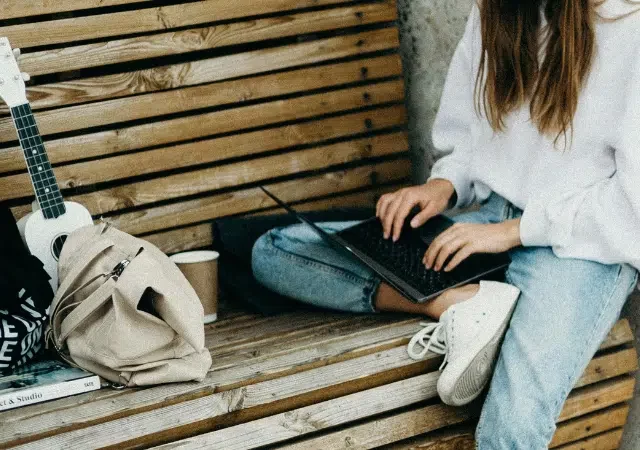 Una persona está sentada en una banca de madera, usando su laptop para trabajar con un creador de correos electrónicos. A su lado hay un ukelele, una mochila beige y un libro, creando un espacio de trabajo sereno lleno de creatividad.