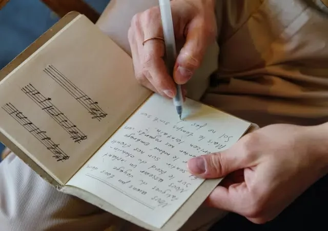 Una persona escribe letras en un cuaderno con bolígrafo; la página del cuaderno a la izquierda muestra notas musicales escritas a mano sobre un pentagrama.