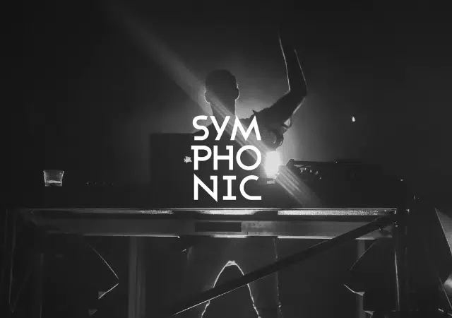 Un DJ está detrás de una mesa con equipos tecnológicos sobre un escenario con iluminación dramática. La palabra “SYMPHONIC” se muestra de forma destacada en letras blancas en negrita.