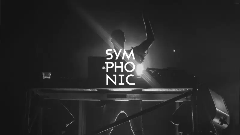 Un DJ está detrás de una mesa con equipos tecnológicos sobre un escenario con iluminación dramática. La palabra “SYMPHONIC” se muestra de forma destacada en letras blancas en negrita.