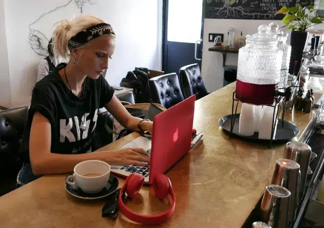 Una persona rubia con una cinta en la cabeza trabaja en una laptop roja sobre una barra de café, con una taza y unos audífonos rojos al lado.