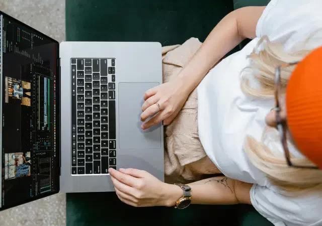 Una persona con gorro naranja y gafas se sienta en un sofá verde usando una laptop, editando metraje de video para una masterclass en línea.