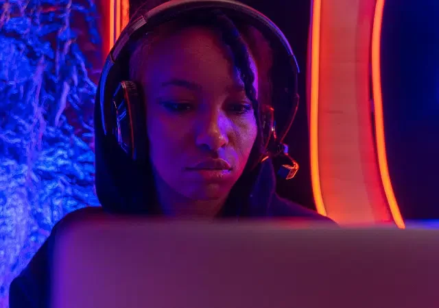 Una persona con auriculares se sienta frente a una laptop, iluminada por luces neón azules y naranjas, inmersa en su trabajo como si actuara para una audiencia invisible.