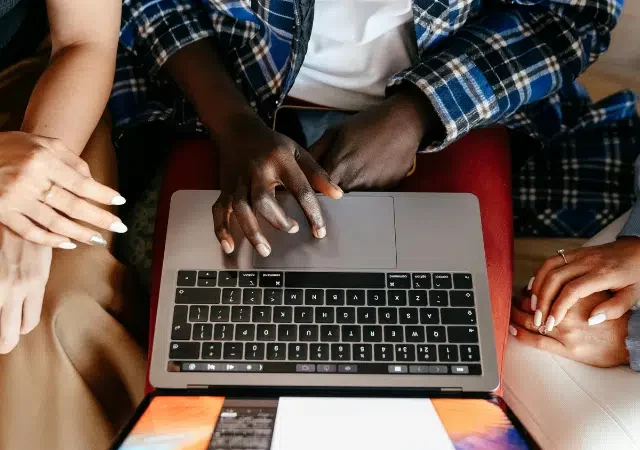 Tres personas sentadas juntas probando algo en una laptop, con sus manos visibles sobre el teclado y el trackpad, vistas desde arriba.
