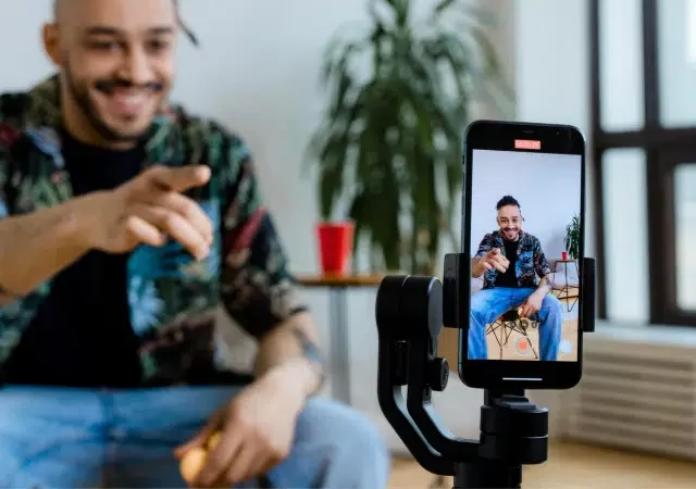 Un hombre está sentado en interiores, sonriendo y señalando un smartphone en un trípode, que lo está grabando. Una planta en maceta y un vaso rojo aparecen en el fondo.