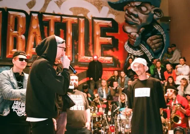 Una batalla de rap tiene lugar frente a una pared con grafitis y una multitud observa; los participantes llevan ropa urbana y gafas de sol.