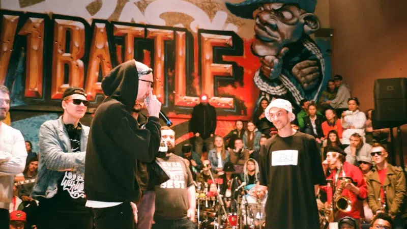 Una batalla de rap tiene lugar frente a una pared con grafitis y una multitud observa; los participantes llevan ropa urbana y gafas de sol.