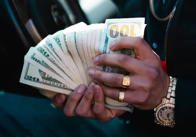 Una persona con joyería dorada, tal vez presumiendo sus regalías, sostiene y abanica un fajo de billetes estadounidenses—principalmente de $20 y $100—dentro de un auto.