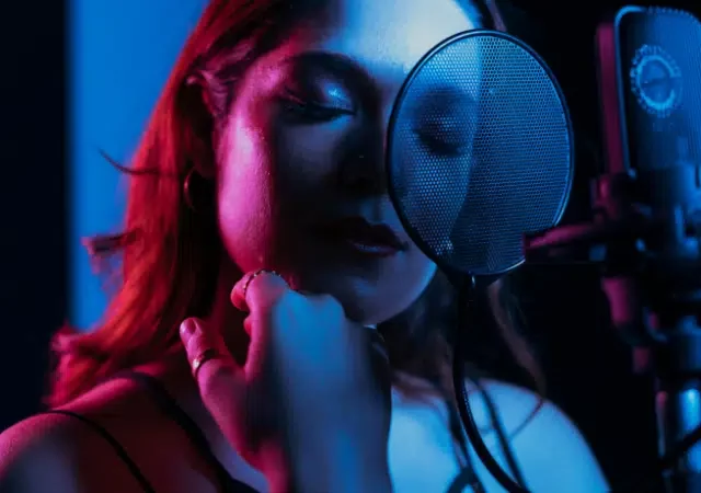 Una mujer frente a un micrófono con filtro pop, cantando o grabando música en un estudio tenuemente iluminado con luces azul y rosa.
