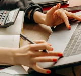 Una persona con esmalte de uñas rojo usa una laptop y escribe en una libreta, con una calculadora sobre el escritorio.