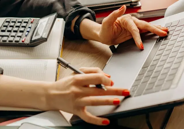 Una persona con esmalte de uñas rojo usa una laptop y escribe en una libreta, con una calculadora sobre el escritorio.