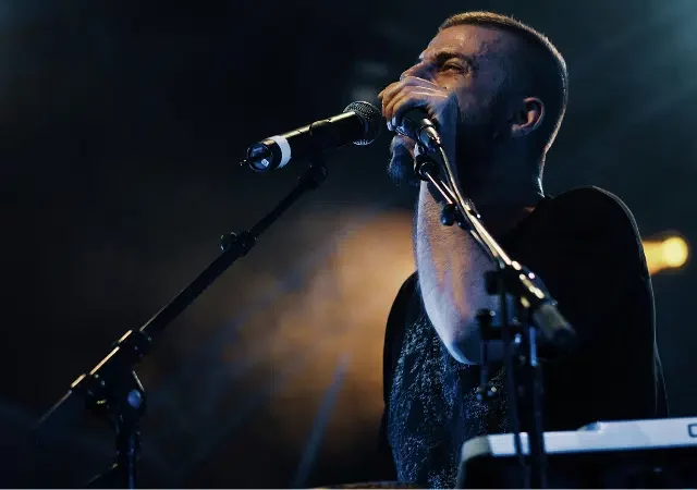 Un hombre está de pie frente a un micrófono en un escenario tenuemente iluminado, cantando o hablando mientras se prepara para el lanzamiento, con un teclado y otro micrófono visibles frente a él.
