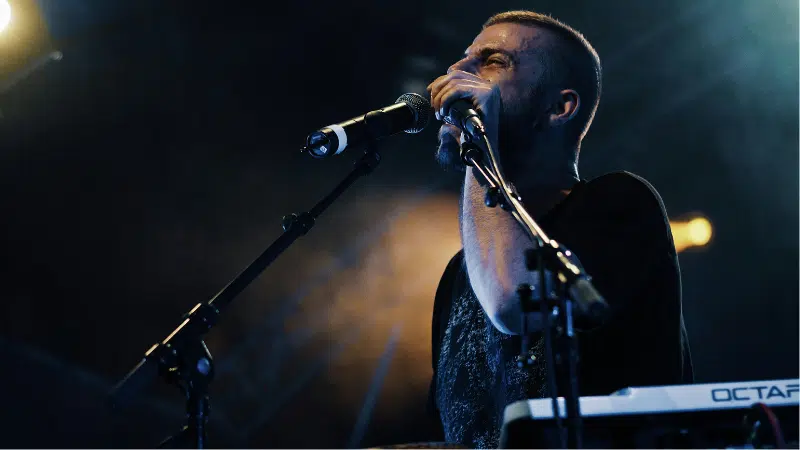 Un hombre está de pie frente a un micrófono en un escenario tenuemente iluminado, cantando o hablando mientras se prepara para el lanzamiento, con un teclado y otro micrófono visibles frente a él.