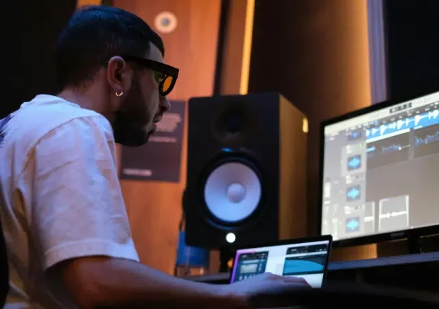 Una persona con gafas se sienta en un escritorio, trabajando en un software de producción musical para su sello en una computadora en un estudio con altavoces e iluminación tenue.