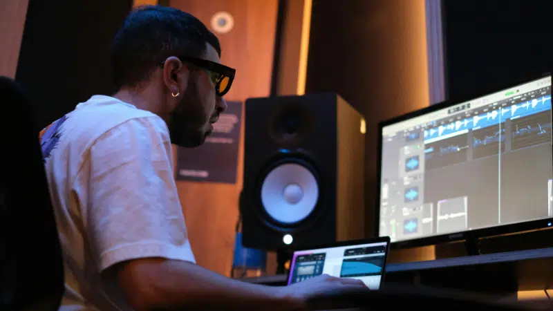 Una persona con gafas se sienta en un escritorio, trabajando en un software de producción musical para su sello en una computadora en un estudio con altavoces e iluminación tenue.