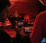 Dos personas graban a una mujer posando en un bar tenuemente iluminado, listas para capturar su mejor look antes del lanzamiento, usando un teléfono inteligente y una cámara con monitor.