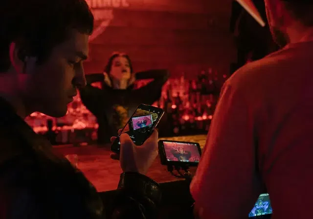 Dos personas graban a una mujer posando en un bar tenuemente iluminado, listas para capturar su mejor look antes del lanzamiento, usando un teléfono inteligente y una cámara con monitor.