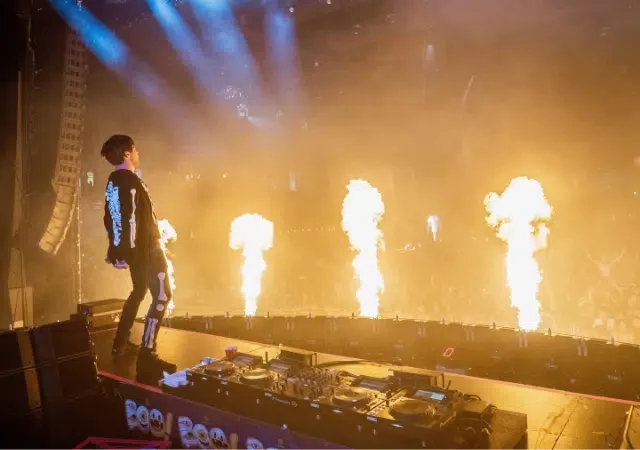 Un DJ se presenta en el escenario con llamas elevándose frente a una multitud de fans enérgicos durante un concierto.