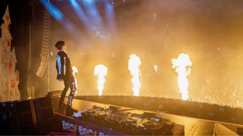 Un DJ se presenta en el escenario con llamas elevándose frente a una multitud de fans enérgicos durante un concierto.