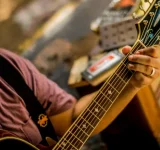 En San Juan, una persona con sombrero y gafas toca una guitarra acústica, concentrándose en formar acordes con la mano izquierda; en el fondo se ve equipo musical.