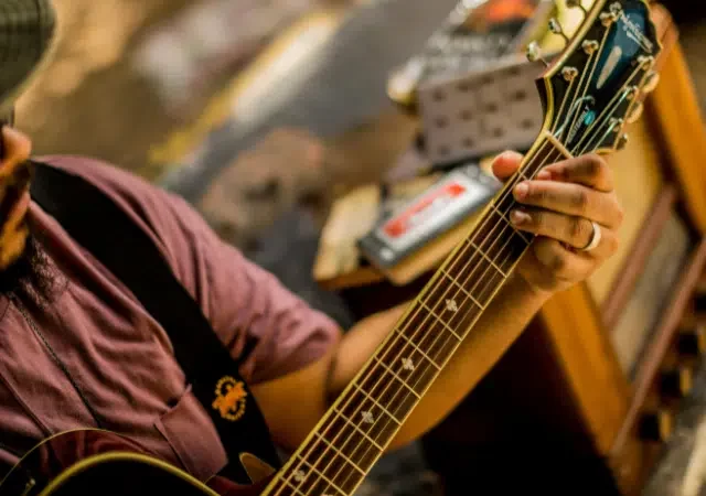 En San Juan, una persona con sombrero y gafas toca una guitarra acústica, concentrándose en formar acordes con la mano izquierda; en el fondo se ve equipo musical.