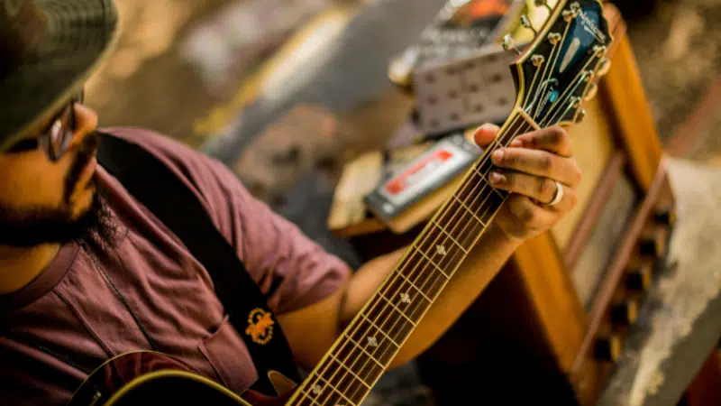 En San Juan, una persona con sombrero y gafas toca una guitarra acústica, concentrándose en formar acordes con la mano izquierda; en el fondo se ve equipo musical.