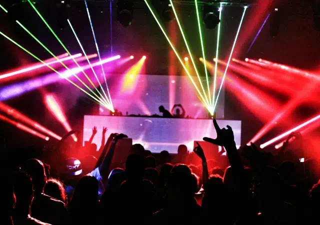 Una multitud frente a la cabina de un DJ en un concierto, mientras luces láser coloridas y humo iluminan el venue oscuro, capturando la vibrante energía de la música en Nueva York.