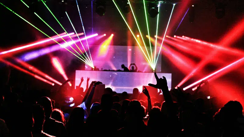 Una multitud frente a la cabina de un DJ en un concierto, mientras luces láser coloridas y humo iluminan el venue oscuro, capturando la vibrante energía de la música en Nueva York.