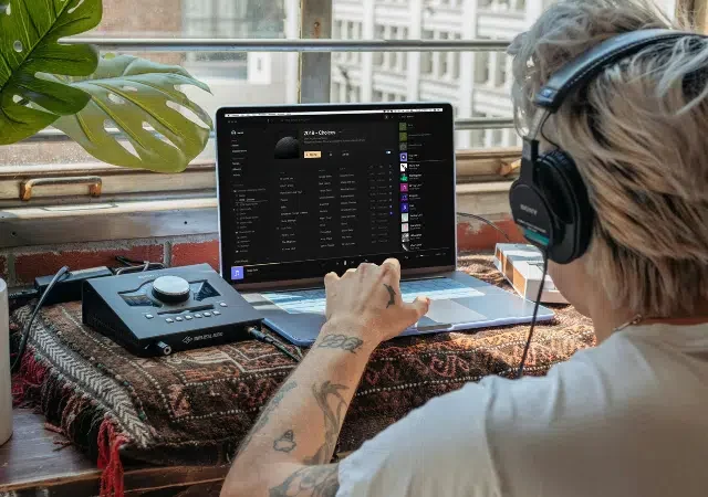 Una persona con audífonos usa una laptop en un escritorio cerca de una ventana, interactuando con equipo de audio y software musical.