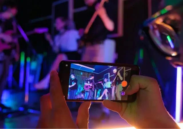 Una persona sostiene un smartphone, grabando o fotografiando a una banda en vivo tocando en el escenario bajo luces coloridas—contenido perfecto para compartir en YouTube for Artists.