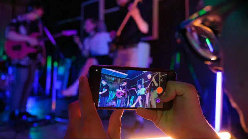 Una persona sostiene un smartphone, grabando o fotografiando a una banda en vivo tocando en el escenario bajo luces coloridas—contenido perfecto para compartir en YouTube for Artists.
