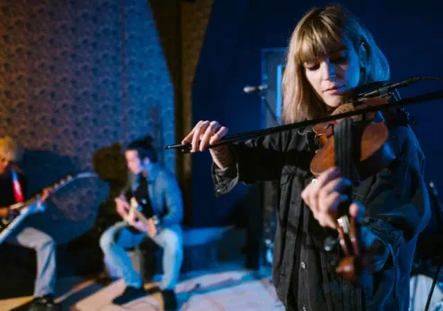 Una mujer toca el violín mientras otros dos músicos tocan guitarras en el fondo, todos interpretando en un espacio interior tenuemente iluminado—mostrando cómo construir un negocio musical escalable a través de presentaciones en vivo cautivadoras.